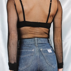 K&ouml;nnte beinhalten: Ein schwarzes, durchsichtiges Langarmshirt mit Stehkragen und einem schwarzen BH darunter. Das Shirt wird mit blauen Jeans mit einem Lee-Logo auf der Ges&auml;&szlig;tasche kombiniert.