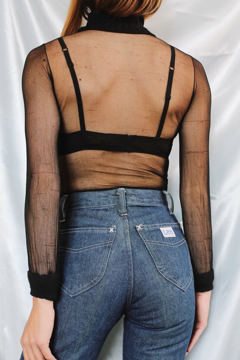 K&ouml;nnte beinhalten: Ein schwarzes, durchsichtiges Langarmshirt mit Stehkragen und einem schwarzen BH darunter. Das Shirt wird mit blauen Jeans mit einem Lee-Logo auf der Ges&auml;&szlig;tasche kombiniert.