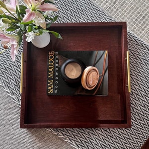 May include: A red mahogany wood tray with gold handles. The tray is on a black and white patterned rug. A book titled "Sam Maloof Woodworker" is on the tray with a candle and a wooden lid.