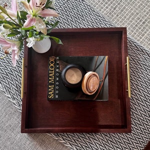 K&ouml;nnte beinhalten: Ein Serviertablett aus rotem Mahagoniholz mit goldenen Griffen. Das Tablett befindet sich auf einer grauen und wei&szlig;en gemusterten Oberfl&auml;che und hat ein Buch mit dem Titel "Sam Malooe Woodworker" darauf.