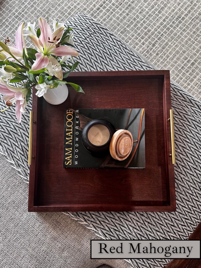 K&ouml;nnte beinhalten: Ein Serviertablett aus rotem Mahagoniholz mit goldenen Griffen. Das Tablett befindet sich auf einer grauen und wei&szlig;en gemusterten Oberfl&auml;che und hat ein Buch mit dem Titel "Sam Malooe Woodworker" darauf.