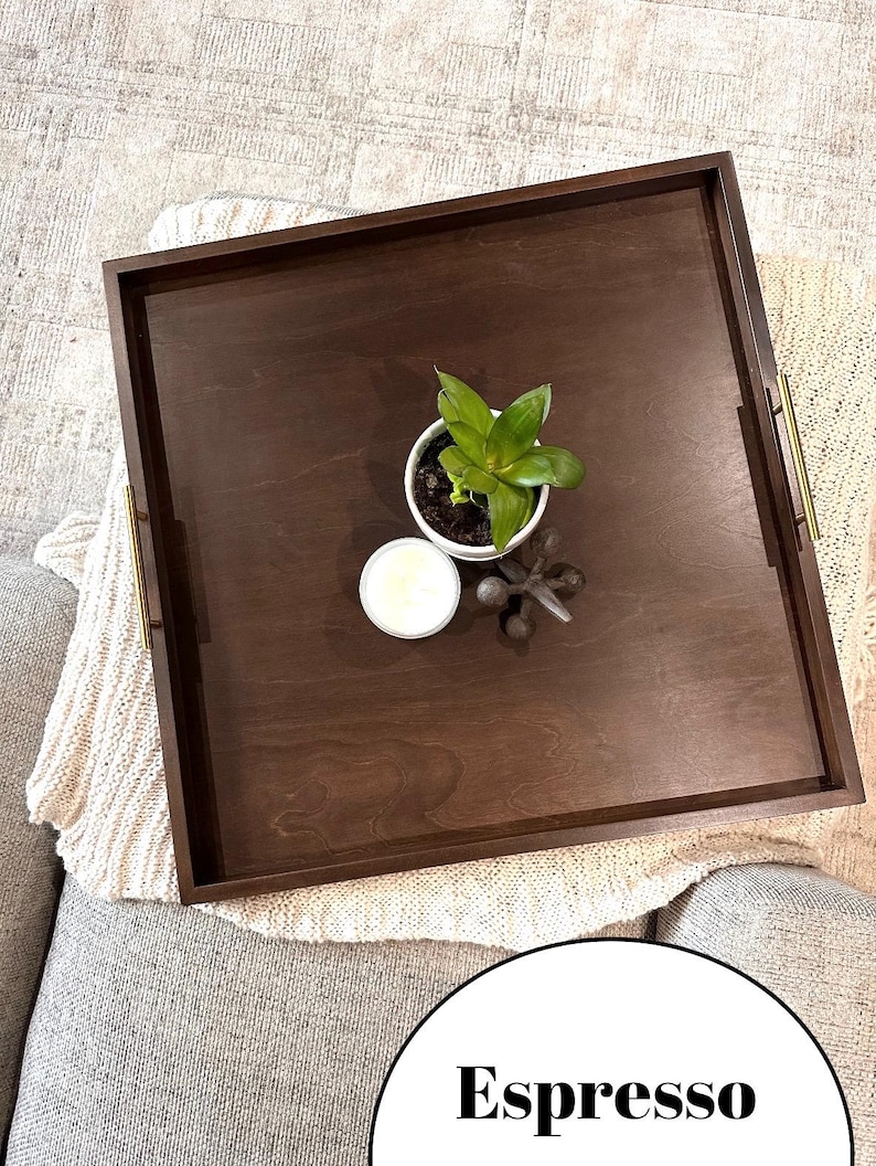 May include: A square wooden tray with brass handles. The tray is espresso brown and has a plant in a white pot, a white candle, and a decorative metal object on top.
