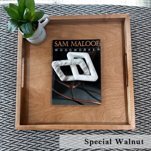May include: A wooden serving tray with a natural wood finish. The tray has two handles on the sides and features the text "SAM MALOOF WOODWORKER" on the surface. The tray is sitting on a black and white patterned surface. The text "Special Walnut" is visible below the tray.