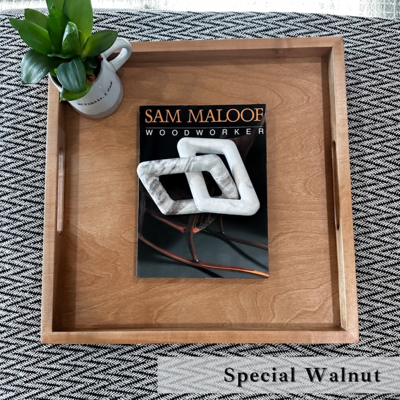 May include: A wooden serving tray with a natural wood finish. The tray has two handles on the sides and features the text "SAM MALOOF WOODWORKER" on the surface. The tray is sitting on a black and white patterned surface. The text "Special Walnut" is visible below the tray.