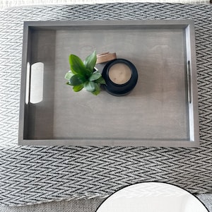 K&ouml;nnte beinhalten: Ein verwittertes graues Holztablett mit einer kleinen Pflanze und einer Kerze in einem schwarzen Glas. Das Tablett hat zwei Griffe an den Seiten.
