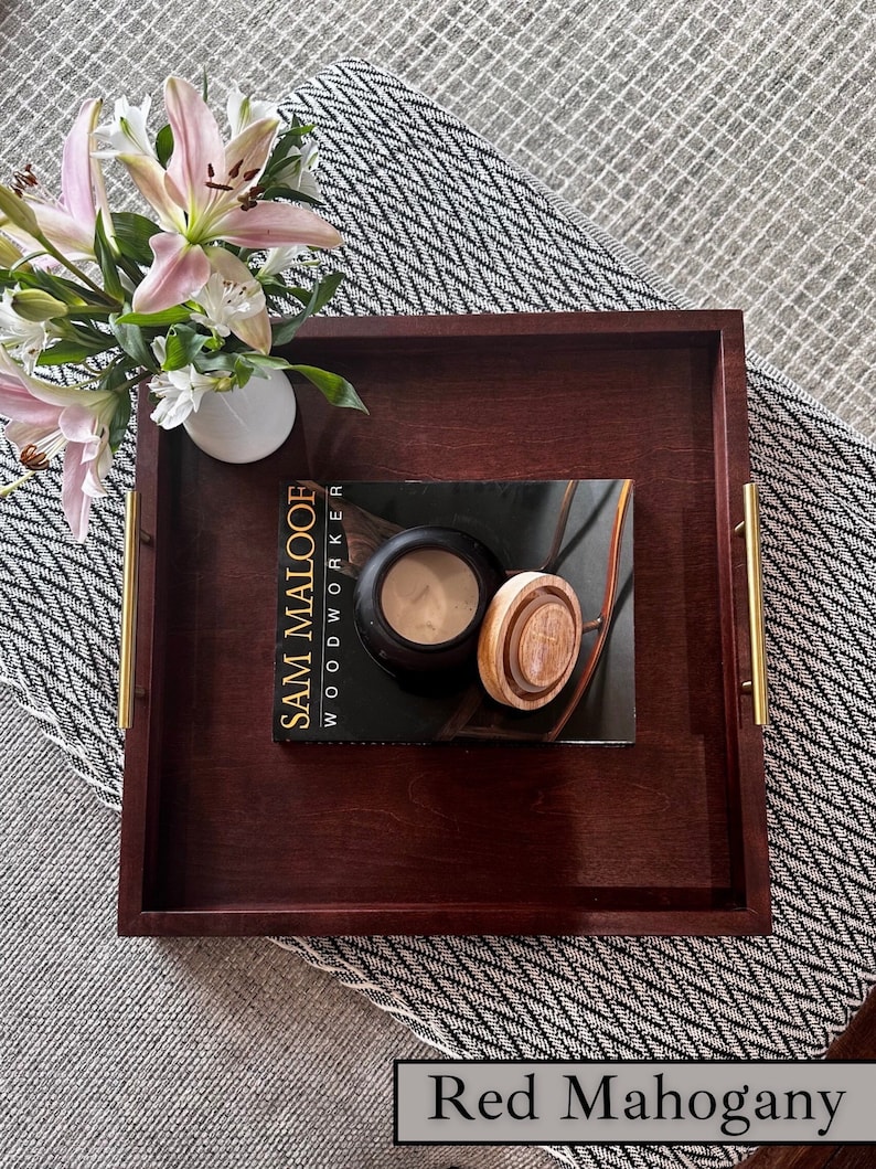 May include: A red mahogany wood tray with gold handles. The tray is on a black and white patterned rug. A book titled "Sam Maloof Woodworker" is on the tray with a candle and a wooden lid.