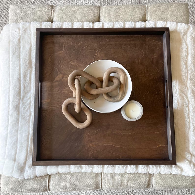 May include: A wooden tray with a dark brown finish. The tray has a white ceramic bowl with a wooden chain inside. A small white candle is on the tray.