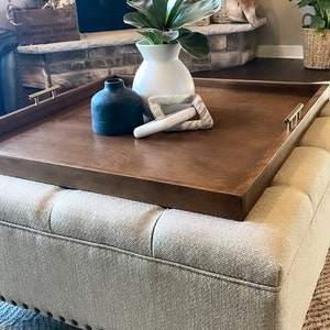 May include: A brown wooden tray with gold handles sits on top of a light gray ottoman with tufted upholstery. The tray is decorated with a white ceramic vase with a green plant, a black ceramic vase, and two white marble decorative pieces.