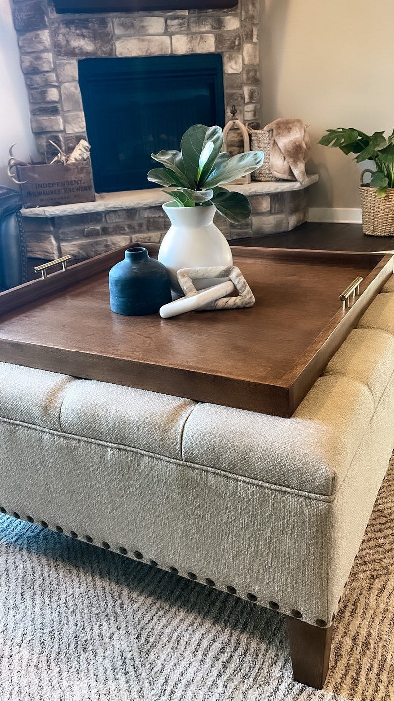 May include: A brown wooden tray with gold handles sits on top of a light gray ottoman with tufted upholstery. The tray is decorated with a white ceramic vase with a green plant, a black ceramic vase, and two white marble decorative pieces.
