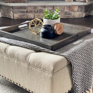 May include: A dark wood tray with a black candle, a gold geometric ornament, a small potted plant, and a wooden lid with the text "Independent Milwaukee Brewery" on a light beige ottoman with a black and white patterned throw blanket.