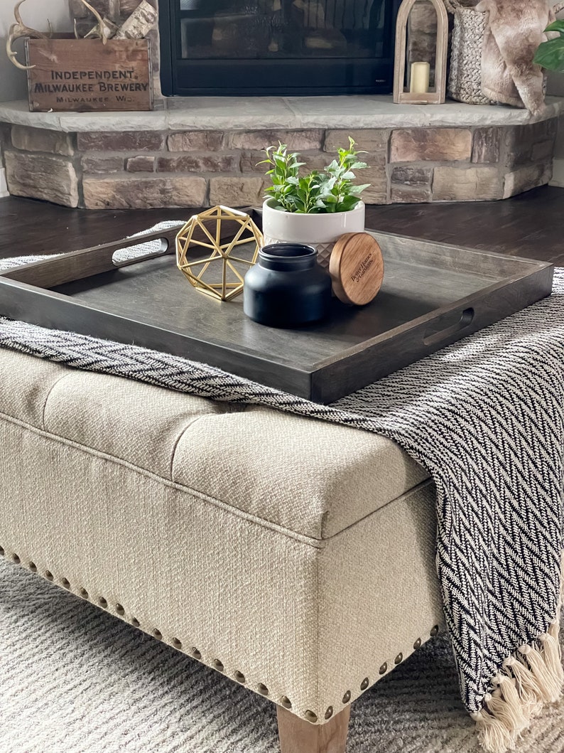 May include: A dark wood tray with a black candle, a gold geometric ornament, a small potted plant, and a wooden lid with the text "Independent Milwaukee Brewery" on a light beige ottoman with a black and white patterned throw blanket.