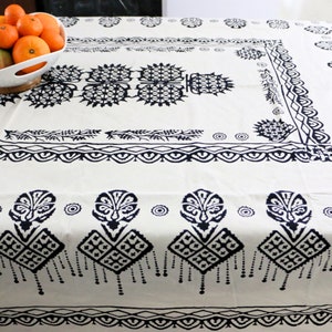 Puede incluir: Un mantel blanco con un patrón geométrico en blanco y negro. El mantel tiene un borde con un diseño repetitivo de hojas estilizadas y un diseño central de una flor estilizada.
