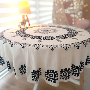 May include: A round, off-white tablecloth with a repeating navy blue geometric pattern. The tablecloth is draped over a white table, with a lamp and decorative horse figurine visible in the background. The tablecloth is made of a soft fabric.