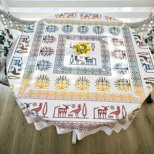 Puede incluir: Un mantel redondo blanco con un colorido estampado geométrico y bordes festoneados. El diseño incluye ciervos, árboles y otras formas en tonos marrones, azules, amarillos y verdes. Una flor amarilla está en el centro de la mesa. Dos cojines florales están en las sillas.