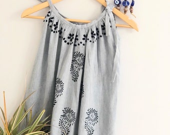 May include: A light blue dress with black floral patterns hangs on a wooden hanger. The dress features tie straps and a round neckline. The floral design is repeated throughout the dress. A decorative wall hanging is visible in the background.