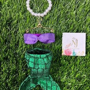 May include: A mermaid-themed set featuring a green and black scale-patterned tail, a purple glittery top, and a pearl bracelet. The set is displayed on a bed of green grass, with a small card that reads "NJ Rejoice".