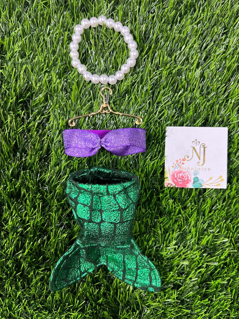 May include: A mermaid-themed set featuring a green and black scale-patterned tail, a purple glittery top, and a pearl bracelet. The set is displayed on a bed of green grass, with a small card that reads "NJ Rejoice".