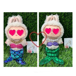 May include: Two plush dolls, each with a bunny hat and mermaid tail. One doll has a pink bow and blue tail, the other has a purple bow and green tail. Both dolls have a pearl necklace and heart-shaped eyes.