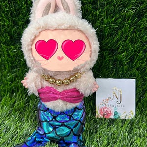 May include: A plush doll dressed as a mermaid bunny. The doll has a fluffy white bunny hood with pink ears, a gold beaded necklace, a pink top, and a blue and green scale-patterned mermaid tail. A small card with the text "Ni&ntilde;aJoice" is visible.