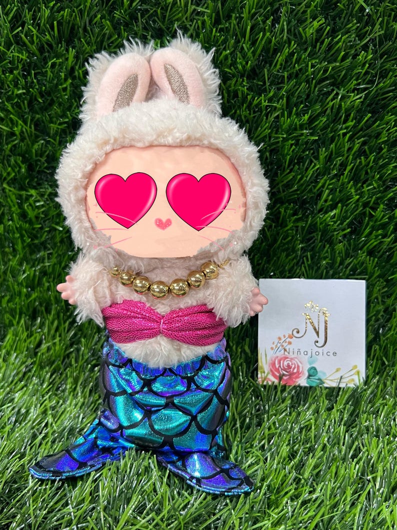 May include: A plush doll dressed as a mermaid bunny. The doll has a fluffy white bunny hood with pink ears, a gold beaded necklace, a pink top, and a blue and green scale-patterned mermaid tail. A small card with the text "Ni&ntilde;aJoice" is visible.