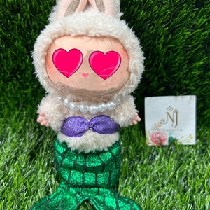 May include: A plush doll dressed as a mermaid with a bunny hood. The doll has a white fluffy hood with bunny ears, a pearl necklace, a purple sequined top, and a green sequined mermaid tail. The doll's eyes are covered with pink hearts.