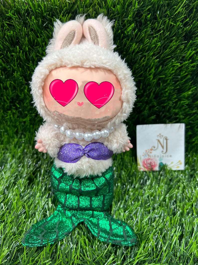 May include: A plush doll dressed as a mermaid with a bunny hood. The doll has a white fluffy hood with bunny ears, a pearl necklace, a purple sequined top, and a green sequined mermaid tail. The doll's eyes are covered with pink hearts.