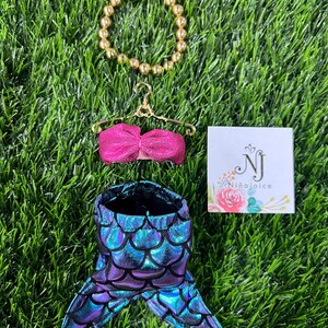 May include: A doll-sized mermaid costume set, including a shimmering blue and purple scale-patterned tail, a pink top, and a gold beaded bracelet. A small card with the text "Ni&ntilde;aJoice" is also present.