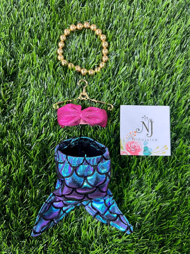 May include: A doll-sized mermaid costume set, including a shimmering blue and purple scale-patterned tail, a pink top, and a gold beaded bracelet. A small card with the text "Ni&ntilde;aJoice" is also present.