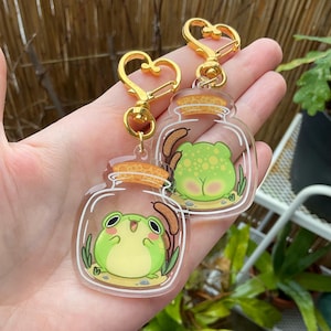 Peut inclure: Deux breloques en forme de bocal en verre transparent avec des porte-clés dorés. Chaque bocal contient une grenouille verte aux yeux noirs. Une grenouille est assise et l'autre est assise dos au spectateur.