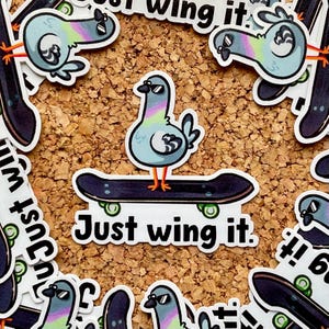 Könnte beinhalten: Ein Aufkleber mit einer Cartoon-Taube, die eine Sonnenbrille trägt und auf einem Skateboard steht. Die Taube ist hellblau, mit regenbogenfarbenen Federn. Das Skateboard ist schwarz mit grünen Rädern, und der Text "Just wing it" steht darunter.