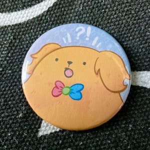 Könnte beinhalten: Ein hellblauer Knopf mit einer Cartoon-Illustration eines goldbraunen Hundes mit einer rosa-blauen Schleife. Der Hund hat einen überraschten Ausdruck und der Text "!?!?" befindet sich über dem Hund.