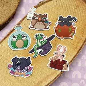 Könnte beinhalten: Satz mit neun bunten Aufklebern mit skurrilen Illustrationen eines Frosches, einer Eule, einer Schlange, einer schwarzen Katze, eines Kaninchens und einer Krähe. Die Aufkleber sind perfekt, um Tagebüchern, Laptops, Wasserflaschen und vielem mehr einen Hauch von Magie zu verleihen.