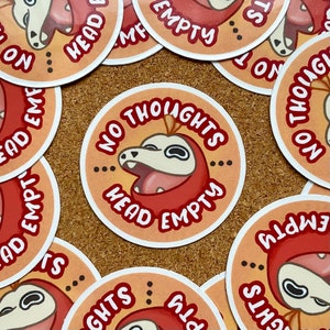 Può includere: Un set di adesivi rotondi con un personaggio dei cartoni animati con la testa rossa e il corpo bianco. Gli adesivi hanno il testo "No Thoughts Head Empty".