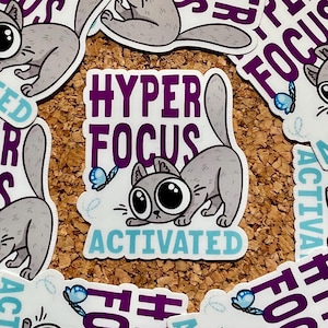 Könnte beinhalten: Ein Aufkleber mit einer Cartoon-grauen Katze mit großen Augen, einem gebogenen Rücken und einem langen Schwanz. Der Aufkleber trägt die Worte "HYPER FOCUS ACTIVATED" in Lila und Türkis. Ein blauer Schmetterling ist ebenfalls abgebildet.