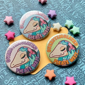 Könnte beinhalten: Drei runde Buttons mit dem Text "OVERSTIMULATED PLEASE WAIT" und einem Cartoon-Kreaturen-Design in Blau, Lila und Grün. Die Buttons sind von sternförmigen Perlen in verschiedenen Farben umgeben, darunter Rosa, Lila und Weiß.