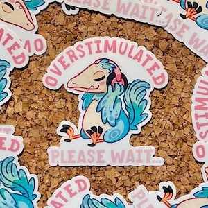 Könnte beinhalten: Eine Sammlung von Aufklebern mit einer Cartoon-Kreatur mit Kopfhörern, beschriftet mit "OVERSTIMULATED PLEASE WAIT...". Die Kreatur ist blau, türkis und beige, mit rosa Akzenten. Die Aufkleber befinden sich auf einer braunen Korkoberfläche.