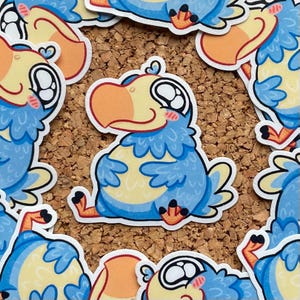 Puede incluir: Una colección de pegatinas de dibujos animados con un pájaro dodo. El dodo tiene plumas azules, vientre amarillo y pico naranja. Las pegatinas están sobre un fondo de corcho.