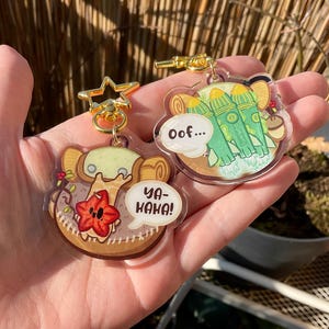 Può includere: Due portachiavi in acrilico con personaggi dei cartoni animati. Un portachiavi presenta una creatura verde con un fiore rosso e il testo "Ya-Haha!" L'altro portachiavi presenta tre razzi verdi con il testo "Oof..."