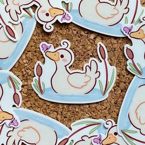 Könnte beinhalten: Weißer Entenaufkleber mit Schmetterling, Schilf und Wassermotiv. Die Ente hat einen gelben Schnabel und eine rosa Wange. Der Aufkleber ist von anderen ähnlichen Aufklebern auf einem Korkbrett-Hintergrund umgeben.