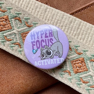 Könnte beinhalten: Ein runder, lila Button mit den Worten "HYPER FOCUS ACTIVATED" in Weiß und einer Cartoon-Katzen-Illustration. Die Katze ist grau und schaut auf einen blauen Schmetterling. Der Button liegt auf einer braunen Oberfläche.