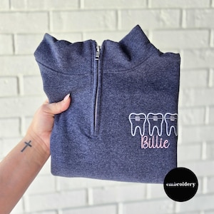 Könnte beinhalten: Marineblaues Sweatshirt mit Viertelreißverschluss, bestickt mit weißen Zähnen und Zahnspangen sowie dem Namen "Billie" in rosa Schrift. Das Sweatshirt hat einen silbernen Reißverschluss und einen hohen Kragen.