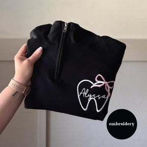 May include: Black quarter-zip sweatshirt with a white embroidered tooth design and a pink bow. The name "Alyssa" is embroidered in cursive within the tooth. The sweatshirt has a silver zipper and the word "embroidery" in a black circle.
