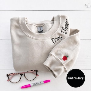 May include: A beige crewneck sweatshirt featuring black embroidered text that reads Mrs. Bloom near the neckline and a small red apple with a green leaf embroidered on the sleeve cuff. A pair of leopard print glasses and a pink marker are included.