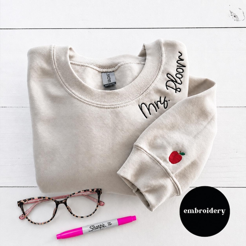 May include: A beige crewneck sweatshirt featuring black embroidered text that reads Mrs. Bloom near the neckline and a small red apple with a green leaf embroidered on the sleeve cuff. A pair of leopard print glasses and a pink marker are included.