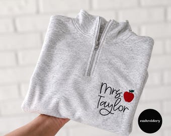 May include: A light gray heathered quarter-zip sweatshirt featuring black embroidered text that reads Mrs. Taylor, accompanied by a small red apple with a green leaf stitched on the right side of the chest near the hem.