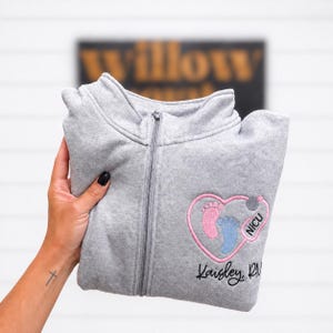 Può includere: Una giacca grigio chiaro con zip caratterizzata da un cuore rosa ricamato contenente un'impronta di piede di neonato rosa e una blu. Il design include un disegno di stetoscopio, il testo NICU e il nome Kaisley RN ricamato in corsivo nero.