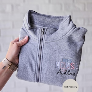 Może przedstawiać: Szara bluza z zamkiem błyskawicznym z haftowanym napisem "Peds Nurse Ashley" i wzorem stetoskopu. Słowo "haft" znajduje się w okręgu poniżej tekstu. Bluza jest złożona i trzymana.