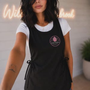 以下が含まれることがあります： 白いTシャツの上に重ねた黒いエプロン。胸元にはカップケーキのグラフィックと「Sweet Creations CUPCAKES」の文字が描かれた円形のピンク色のロゴがあしらわれています。サイドで結ぶ紐が付いた、シンプルで機能的なデザインのエプロンです。