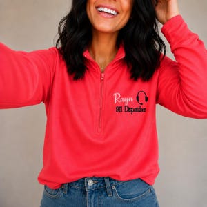 May include: A bright coral long-sleeve quarter-zip pullover featuring black embroidery on the left chest. The text reads Rayn followed by 911 Dispatcher with a small black headset icon. The garment has a high collar and a vertical zipper closure.