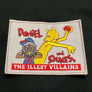 May include: A grey embroidered patch featuring a yellow cartoon character and a red and blue cartoon character. The text on the patch reads "Daniel and Quas, The Illest Villains".